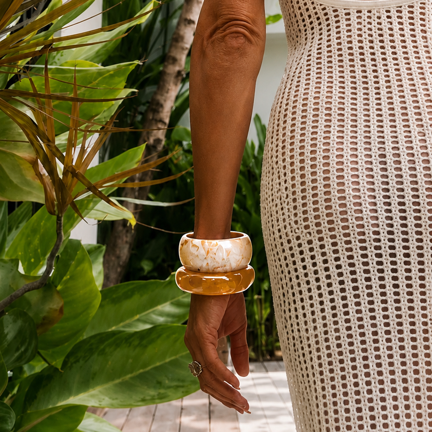 A woman viewed from behind wearing a cream open-knit crochet maxi dress with thin straps, standing on a wooden deck surrounded by lush tropical greenery.
