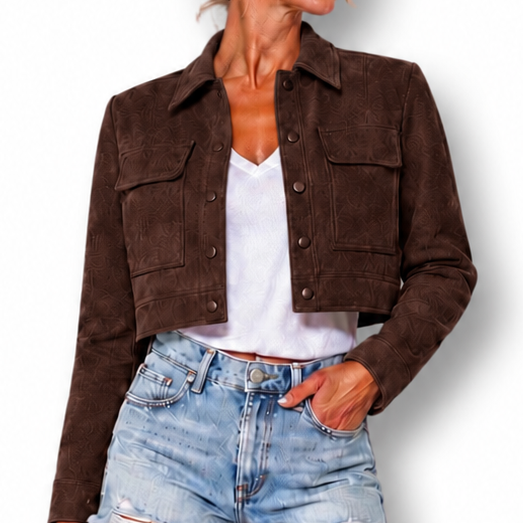A woman modeling a dark brown cropped suede jacket over a white top and light-wash denim shorts, standing against a plain white background.