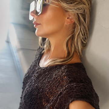 A woman wearing a dark brown open-knit short-sleeve top and clear-framed sunglasses, posing in a selfie-style shot against a light-colored wall.