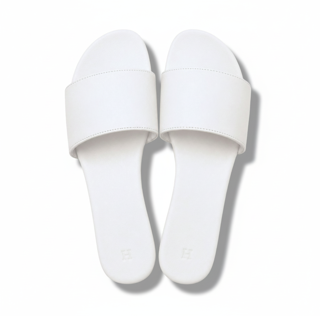 A pair of minimalist white slide sandals with wide, flat straps, shown from a top-down view on a clean white background.