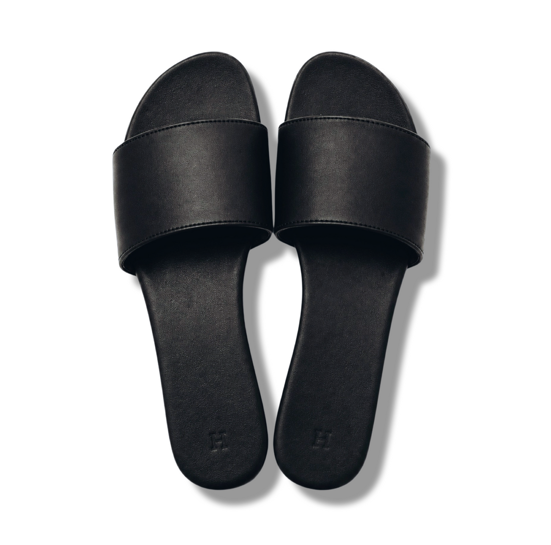 A pair of black slide sandals with wide, flat straps and a minimalist design, photographed from a top-down angle on a white background.