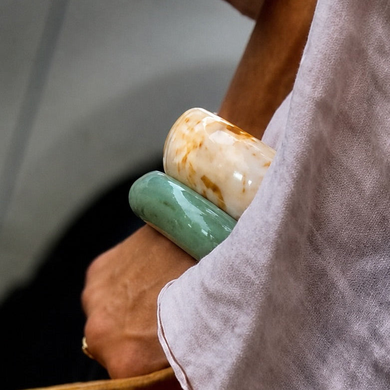 A close-up lifestyle shot of a person wearing a textured light gray linen shirt and holding a brown leather bag, accessorized with a large green and cream stone bangle.